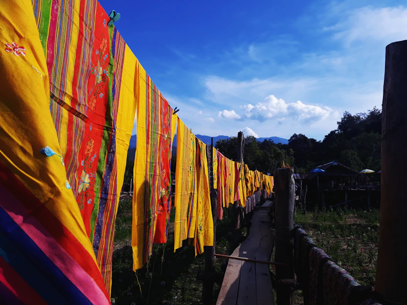 A serene riverside tribal village with handcrafted textiles drying in the sun and artisans weaving under tall trees.