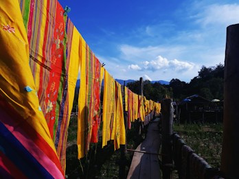 Brightly colored textiles with intricate patterns are hung on a line against a backdrop of clear blue skies and distant mountains. The textiles feature vibrant yellows, reds, and greens with floral and striped designs. A wooden pathway is visible below the textiles, and trees fill the surrounding landscape.