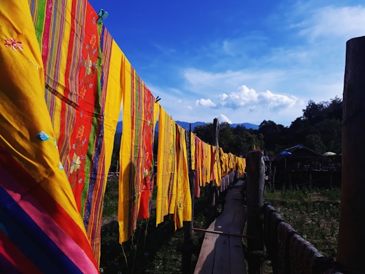 Brightly colored textiles with intricate patterns are hung on a line against a backdrop of clear blue skies and distant mountains. The textiles feature vibrant yellows, reds, and greens with floral and striped designs. A wooden pathway is visible below the textiles, and trees fill the surrounding landscape.