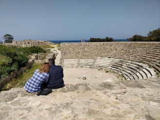 Two people sitting closely on a stone terrace, overlooking a partially ruined ancient amphitheater. The scene is set in a historical site with lush greenery and a view of the sea in the background.