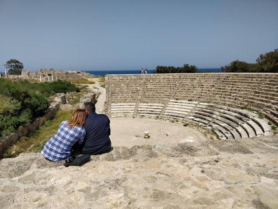 Two people sitting closely on a stone terrace, overlooking a partially ruined ancient amphitheater. The scene is set in a historical site with lush greenery and a view of the sea in the background.