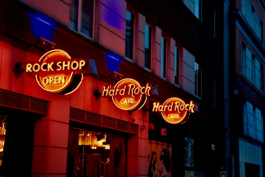 A city street scene features a prominent building facade with bright, neon signs for a Rock Shop and Hard Rock Cafe. The signs are illuminated in orange and yellow hues, casting a warm glow against the dark exterior of the building. The windows on the building reflect some ambient light, while the street remains dimly lit, enhancing the vibrant contrast.