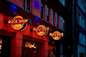 A city street scene features a prominent building facade with bright, neon signs for a Rock Shop and Hard Rock Cafe. The signs are illuminated in orange and yellow hues, casting a warm glow against the dark exterior of the building. The windows on the building reflect some ambient light, while the street remains dimly lit, enhancing the vibrant contrast.