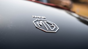 A close-up of an MG car badge on a glossy black surface, reflecting light softly. The badge is prominently silver with a geometric design, surrounded by a blurred background.