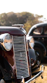 A vintage car with a glossy, dark red finish and prominent chrome details, including a large grille and round headlights. The background shows another classic vehicle and blurred trees, suggesting an outdoor setting.