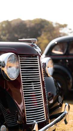 A vintage car with a glossy, dark red finish and prominent chrome details, including a large grille and round headlights. The background shows another classic vehicle and blurred trees, suggesting an outdoor setting.