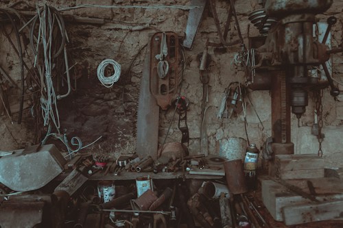 A cluttered and rustic workshop filled with various tools and equipment. The walls are made of rough stone, and a variety of hanging tools and wires are organized chaotically. A large drill press is visible on the right, surrounded by cans and metal scraps. The atmosphere is dusty and aged, with muted earthy colors throughout.