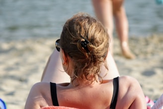 A person with brown wavy hair is lounging on a sandy beach. They are wearing sunglasses and a black swimsuit, holding a book. Another person is walking towards the water in the background.