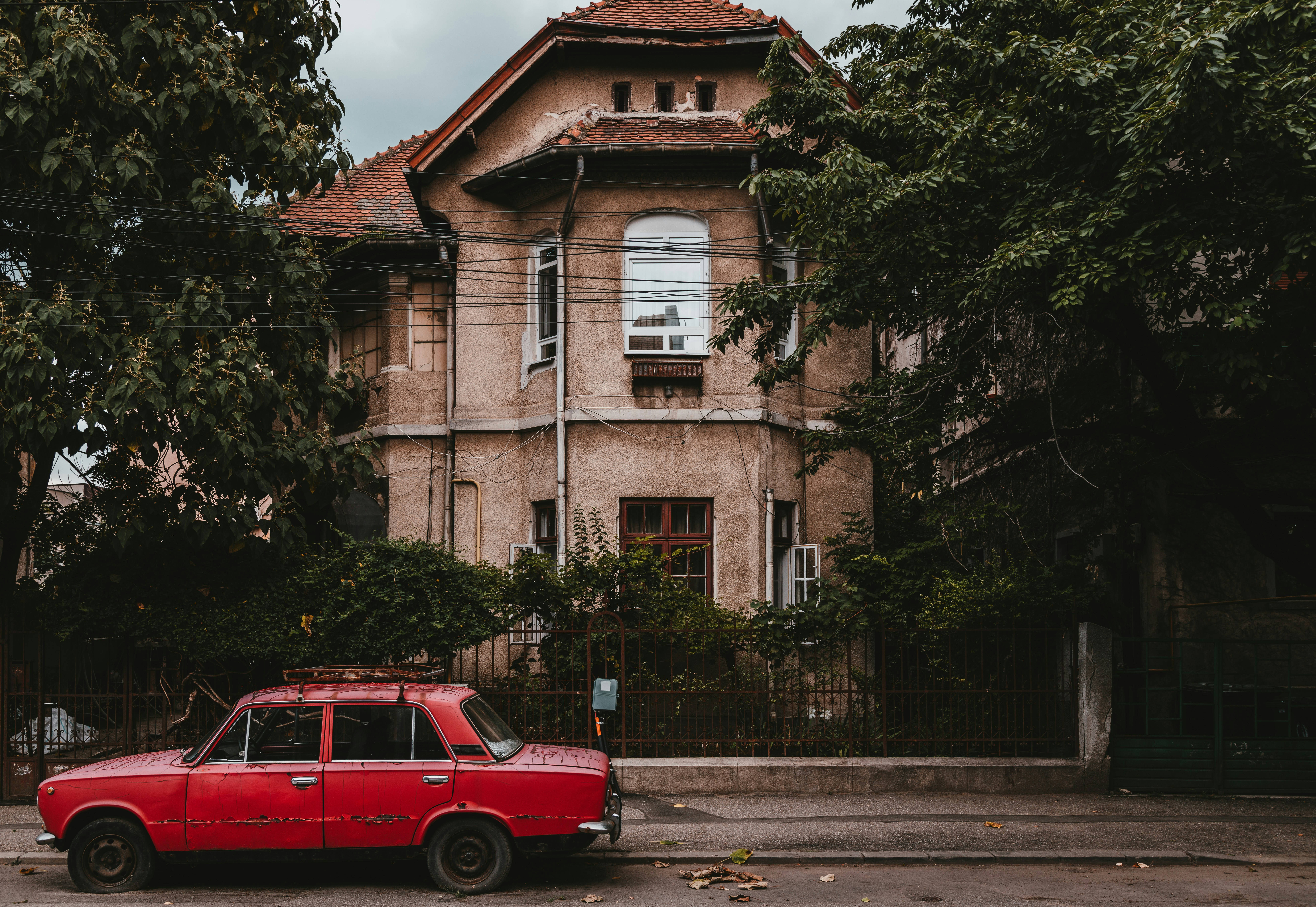 red sedan parking near road beside house surrounded with tall and green trees