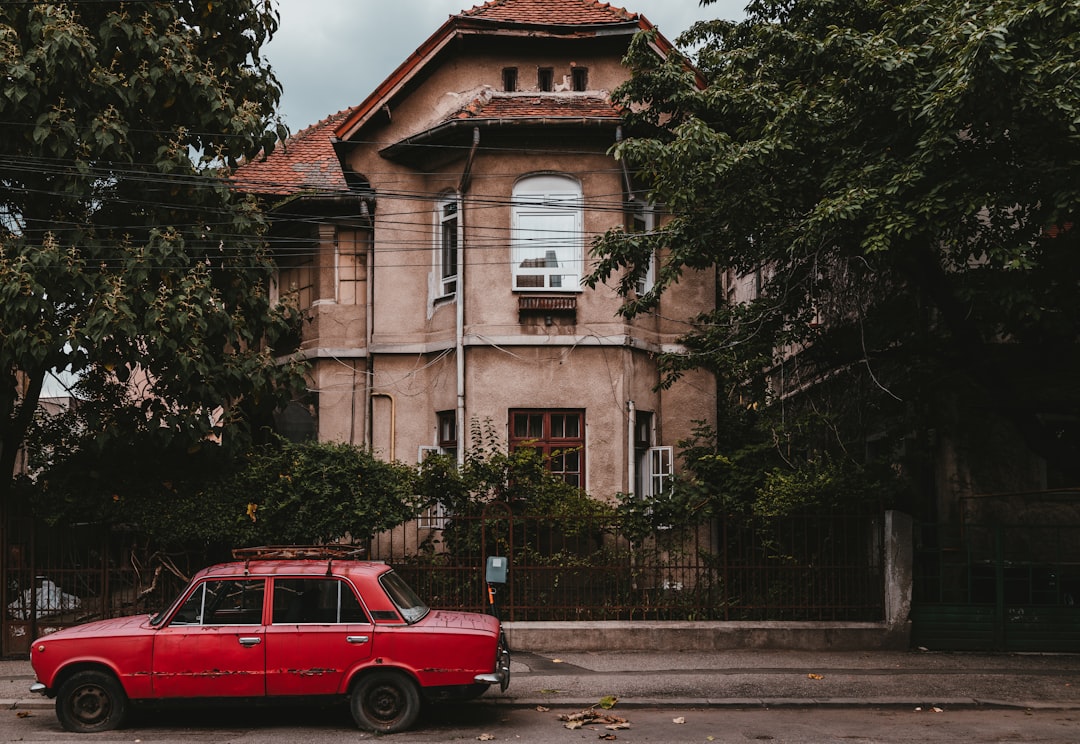 red sedan parking near road beside house surrounded with tall and green trees