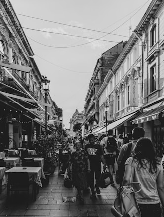 A bustling urban street scene with people walking along a narrow pedestrian path lined with cafes. The architecture is European, with ornate buildings and streetlamps, and the atmosphere is lively and social. The photograph is in black and white, adding a classic, timeless feel.