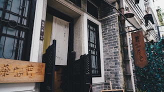 A rustic, traditional tea house entrance with a wooden sign featuring Chinese characters is flanked by stone walls and decorative black metal grills. A fabric banner with subtle calligraphy adds a cultural touch, while hanging lanterns and greenery create a cozy, inviting atmosphere.
