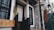 A rustic, traditional tea house entrance with a wooden sign featuring Chinese characters is flanked by stone walls and decorative black metal grills. A fabric banner with subtle calligraphy adds a cultural touch, while hanging lanterns and greenery create a cozy, inviting atmosphere.
