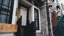A rustic, traditional tea house entrance with a wooden sign featuring Chinese characters is flanked by stone walls and decorative black metal grills. A fabric banner with subtle calligraphy adds a cultural touch, while hanging lanterns and greenery create a cozy, inviting atmosphere.