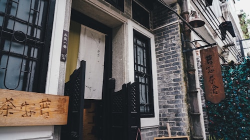A rustic, traditional tea house entrance with a wooden sign featuring Chinese characters is flanked by stone walls and decorative black metal grills. A fabric banner with subtle calligraphy adds a cultural touch, while hanging lanterns and greenery create a cozy, inviting atmosphere.