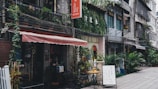 A quaint street scene features a series of shops and cafes with a charming facade. Plants and greenery climb the building's exterior, adding a touch of nature. A sign with writing in a foreign language hangs above, and a chalkboard stands outside with text displayed. Tables and chairs are set up, inviting passersby to sit.