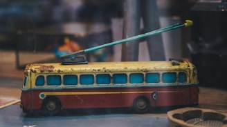 A vintage toy bus with a yellow roof, red body, and blue windows is shown with a long metal rod attachment. The toy appears to be made of metal with visible rust and wear, indicating its age. The background includes blurred objects and colors, giving the impression of a retro setting.