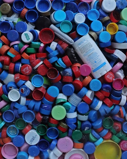 A collection of various colored plastic bottle caps and lids, ranging in colors from blue, red, green, pink, to orange, are packed together. Among them, a cylindrical plastic container with text is visible.