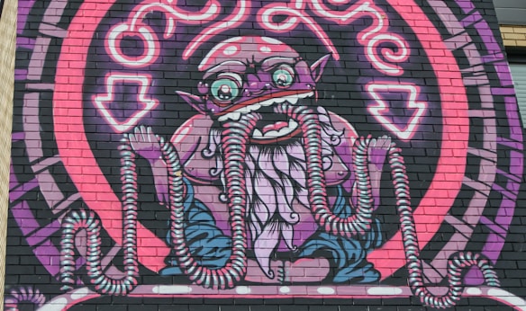 An intricate mural on a brick wall features a fantastical creature with exaggerated features, including large eyes, pointed ears, and multiple tentacles extending from its face and hands. The creature is surrounded by a circular, neon-like pattern with vibrant shades.