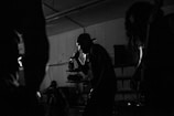 Silhouettes of musicians performing in a dimly lit space, including a central figure holding a microphone and singing passionately. The background reveals shelves and equipment, contributing to the atmosphere of a live music venue.