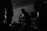 Silhouettes of musicians performing in a dimly lit space, including a central figure holding a microphone and singing passionately. The background reveals shelves and equipment, contributing to the atmosphere of a live music venue.