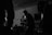 Silhouettes of musicians performing in a dimly lit space, including a central figure holding a microphone and singing passionately. The background reveals shelves and equipment, contributing to the atmosphere of a live music venue.