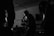 Silhouettes of musicians performing in a dimly lit space, including a central figure holding a microphone and singing passionately. The background reveals shelves and equipment, contributing to the atmosphere of a live music venue.