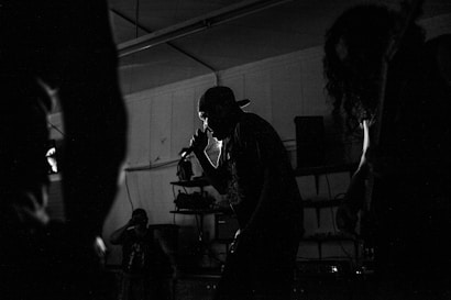 Silhouettes of musicians performing in a dimly lit space, including a central figure holding a microphone and singing passionately. The background reveals shelves and equipment, contributing to the atmosphere of a live music venue.