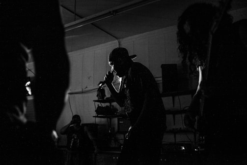 Silhouettes of musicians performing in a dimly lit space, including a central figure holding a microphone and singing passionately. The background reveals shelves and equipment, contributing to the atmosphere of a live music venue.