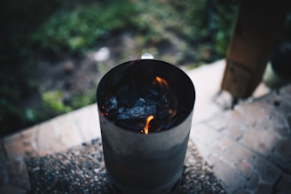 A charcoal chimney starter with visible flames is positioned on a gritty surface, likely a patio. The background shows a blurred grassy area and a wooden post, giving a hint of an outdoor setting.