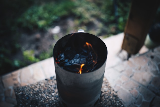 A charcoal chimney starter with visible flames is positioned on a gritty surface, likely a patio. The background shows a blurred grassy area and a wooden post, giving a hint of an outdoor setting.