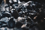 A close-up view of several pieces of coal or charcoal with rough and uneven textures. The image focuses on the dark, black tones of the material, with some highlights catching the light, creating a stark contrast against the shadows.
