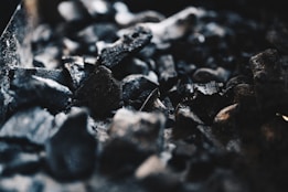 A close-up view of several pieces of coal or charcoal with rough and uneven textures. The image focuses on the dark, black tones of the material, with some highlights catching the light, creating a stark contrast against the shadows.