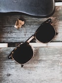 A pair of dark sunglasses rests on a worn wooden surface. Near the sunglasses, there is a small, dried leaf. A black, textured eyeglass case also lies on the surface.