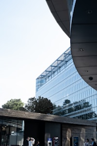 A modern architectural scene with a large glass building reflecting trees and sky, adjacent to a smaller structure with a dark wooden facade. A few people are visible near the entrance and contemporary design elements.
