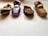 Four ballet pointe shoes are arranged on a flat surface in a row. The shoes, differing in color and wear, include a shiny purple pair alongside beige and brown ones. Each shoe displays signs of use, with creased fabric and worn tips.