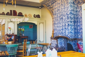 A cozy vintage-style room with a strong Dutch aesthetic is adorned with intricate blue and white Delft tiles covering a large portion of the wall and fireplace. Wooden furniture, including chairs and tables, is arranged throughout the space. A retro television and other decorative items sit on the shelves and tables, contributing to the room's nostalgic feel. The room is warmly lit, enhancing the antique and charming atmosphere.