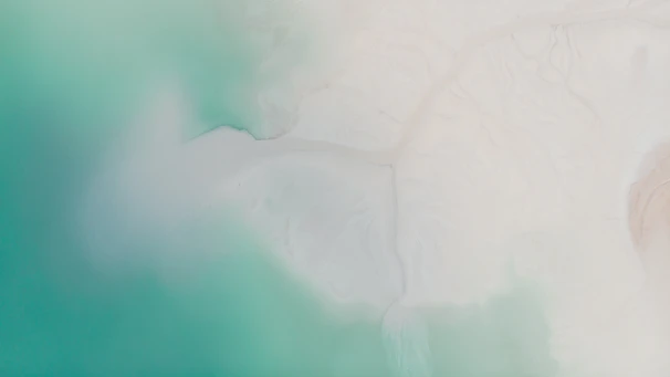 Aerial watercolor illustration of turquoise sea meeting warm sandy beach with lush green tropical foliage.