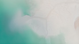 Aerial view of a coastline where vibrant turquoise waters meet a sandy, white shore. The blending of colors creates a serene atmosphere with soft gradients and smooth textures.
