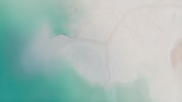 Aerial view of a coastline where vibrant turquoise waters meet a sandy, white shore. The blending of colors creates a serene atmosphere with soft gradients and smooth textures.