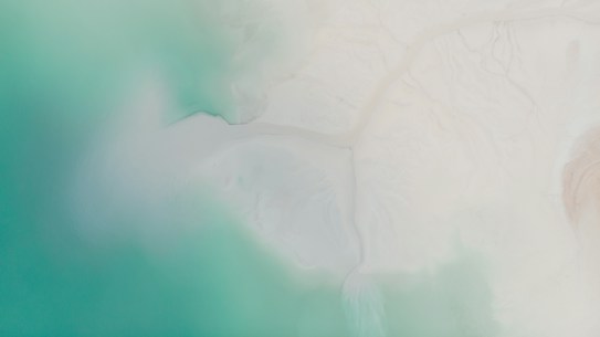 Aerial view of a coastline where vibrant turquoise waters meet a sandy, white shore. The blending of colors creates a serene atmosphere with soft gradients and smooth textures.