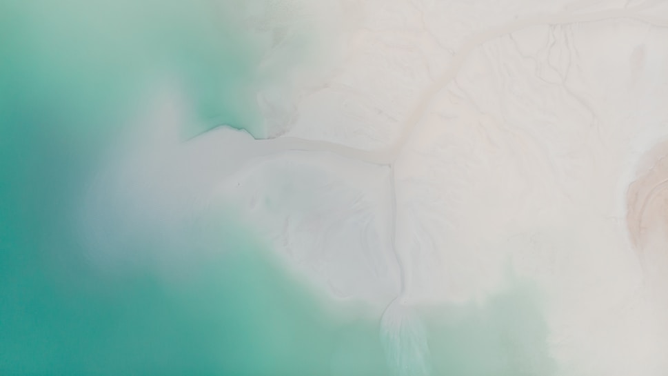 Aerial view of a coastline where vibrant turquoise waters meet a sandy, white shore. The blending of colors creates a serene atmosphere with soft gradients and smooth textures.