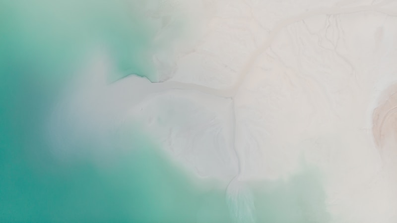 Aerial view of a coastline where vibrant turquoise waters meet a sandy, white shore. The blending of colors creates a serene atmosphere with soft gradients and smooth textures.