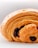 A close-up view of a golden-brown pastry with flaky layers and a visible chocolate filling. The pastry shows intricate, delicate layers and looks crispy on the outside. The background is blurred and white, putting focus on the pastry.