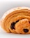 A close-up view of a golden-brown pastry with flaky layers and a visible chocolate filling. The pastry shows intricate, delicate layers and looks crispy on the outside. The background is blurred and white, putting focus on the pastry.