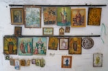 A collection of various religious icons and paintings adorns a wall, featuring different figures and scenes, mostly painted in vibrant colors. Some of the icons are framed, while others hang unframed. The artworks depict saints, biblical figures, and religious scenes, with intricate details and a mix of artistic styles.