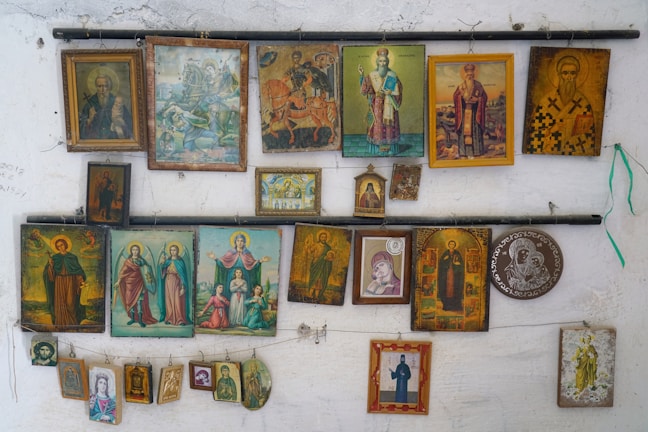 A collection of various religious icons and paintings adorns a wall, featuring different figures and scenes, mostly painted in vibrant colors. Some of the icons are framed, while others hang unframed. The artworks depict saints, biblical figures, and religious scenes, with intricate details and a mix of artistic styles.