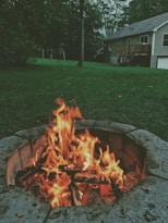 A campfire burning brightly in a stone fire pit, surrounded by lush grass. In the background, a house with a wooden deck sits amidst tall trees. The sky appears overcast, suggesting a calm dusk or early evening setting.