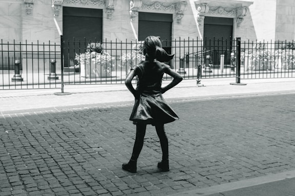 A bronze statue of a young girl with her hands on her hips stands confidently on a cobblestone street. She faces a building with ornate architecture and closed shutters, separated by a black iron fence. The image conveys a sense of empowerment through the girl's determined stance.