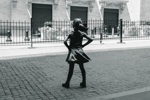 A bronze statue of a young girl with her hands on her hips stands confidently on a cobblestone street. She faces a building with ornate architecture and closed shutters, separated by a black iron fence. The image conveys a sense of empowerment through the girl's determined stance.